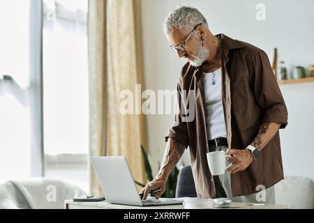 Un homme mature et tatoué aux cheveux gris travaille sur son ordinateur portable tout en tenant une tasse de café. Banque D'Images