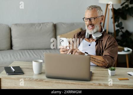 Un homme gay mature avec des tatouages et des cheveux gris travaille à distance de chez lui, souriant en regardant son téléphone. Banque D'Images