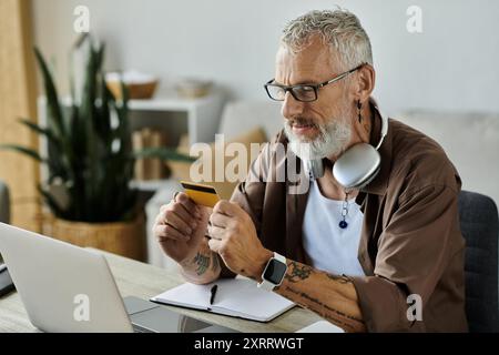 Un homme gay mature avec des tatouages et des cheveux gris est assis à son bureau travaillant à distance à la maison, tenant une carte de crédit et regardant son ordinateur portable. Banque D'Images