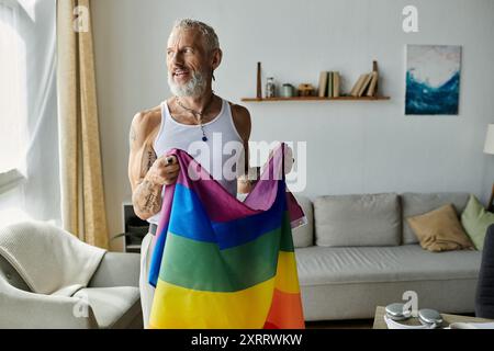 Un homme gay mature avec des tatouages et des cheveux gris sourit et tient un drapeau arc-en-ciel de fierté dans son salon. Banque D'Images