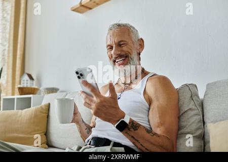 Un homme mûr avec des tatouages et une barbe grise sourit alors qu'il se détend sur son canapé à la maison, faisant défiler son téléphone. Banque D'Images