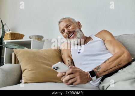 Un homme gay mature avec des tatouages et une barbe grise se détend sur un canapé à la maison, souriant et tenant un smartphone. Banque D'Images