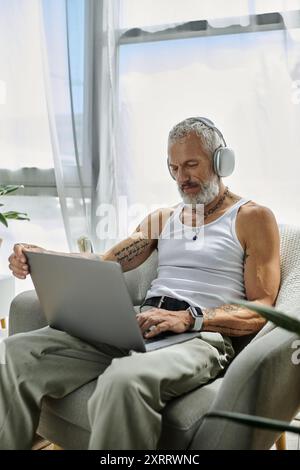 Un homme tatoué à la barbe grise se détend sur son canapé tout en utilisant un ordinateur portable. Banque D'Images