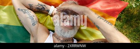 Un homme gay mature avec des tatouages et une barbe grise gît sur un drapeau arc-en-ciel à l'extérieur, souriant. Banque D'Images