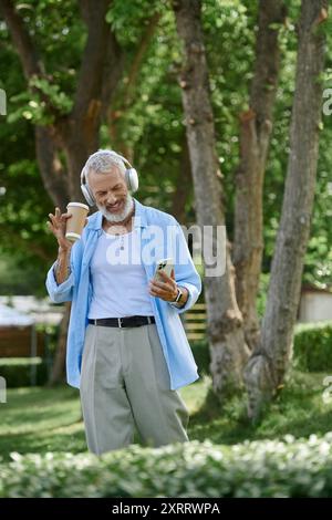 Homme avec des tatouages, barbe grise profite d'une journée ensoleillée dans le parc, écoutant de la musique avec du café. Banque D'Images