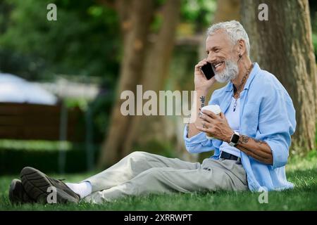 Un homme gay mature avec des tatouages et une barbe grise est assis sur l'herbe, souriant tout en parlant sur son téléphone. Banque D'Images