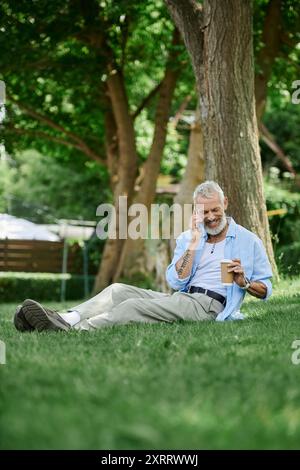 Un homme gay mature avec des tatouages et une barbe grise est assis sur l'herbe à l'extérieur. Il est au téléphone et prend un café. Banque D'Images