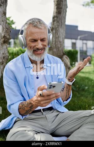 Un homme gay mature avec des tatouages et une barbe grise est assis sur l'herbe à l'extérieur, portant des écouteurs et souriant en regardant son téléphone. Banque D'Images