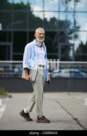 Un homme gay mûr avec une barbe grise et des tatouages se promène dehors avec un ordinateur portable et des écouteurs autour du cou. Banque D'Images