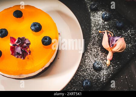 Dessert ou gâteau avec des couches de chocolat, crème caillée, gelée de mangue, décoré de fleurs séchées sur une assiette blanche sur fond noir. Original Banque D'Images