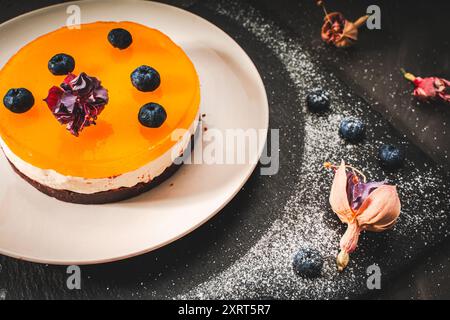 Dessert ou gâteau avec des couches de chocolat, crème caillée, gelée de mangue, décoré de fleurs séchées sur une assiette blanche sur fond noir. Original Banque D'Images