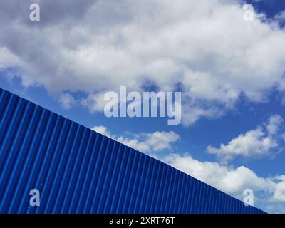 Plaque métallique bleue contre ciel nuageux bleu. Bardage. Surface sans soudure en acier galvanisé. Mur de bâtiment industriel en tôle ondulée, plat Banque D'Images