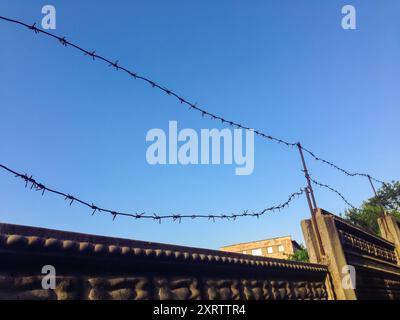 Une clôture en fil de fer avec du fil de fer barbelé est suspendue au-dessus d'un mur. Le ciel est clair et bleu Banque D'Images