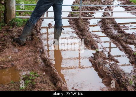 Des pistes boueuses, pleines d'eau et ornés par des véhicules passant par une porte de ferme où un homme se tient debout dans une paire de bottes wellington. Banque D'Images