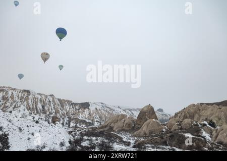 Montgolfières flottant au-dessus du paysage enneigé de la Cappadoce, mettant en valeur un terrain rocheux et des cheminées de fées pendant un matin d'hiver serein Banque D'Images