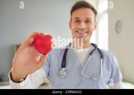 Médecin cardiologue souriant tenant le cœur rouge dans les mains et regardant la caméra dans le cabinet médical Banque D'Images