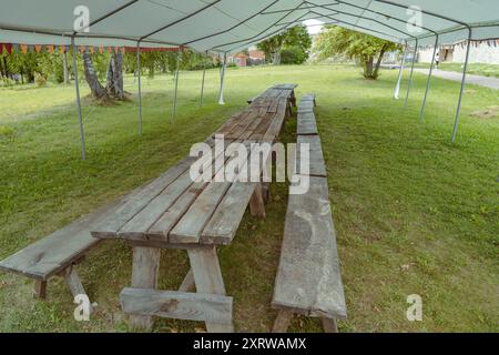 Des tables en bois rustiques sont placées sous une grande tente dans une zone herbeuse. Banque D'Images