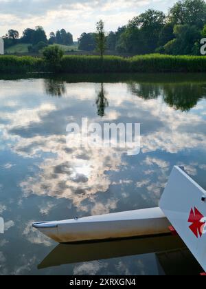 Fondé en 1921 et situé sur une belle partie de la Tamise dans l'Oxfordshire, Radley Boathouse sert Radley College et les amateurs d'aviron locaux depuis plus d'un siècle. Si vous vous tenez sur sa jetée et regardez à travers la rive nord de la rivière, vous verrez un arbre isolé et plutôt charmant, qui semble changer de caractère et d'apparence tout au long de l'année. Ici, nous le voyons un matin d'été nuageux, avec les catamarans de plaisance omniprésents du hangar à bateaux au premier plan. La Croix de Malte rouge est l'emblème du club de navigation de l'université. Banque D'Images