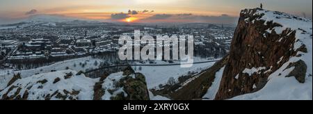 Vue panoramique d'Édimbourg, en Écosse, couverte de neige depuis Arthur Seat, avec le coucher du soleil derrière la ville et une personne debout sur le bord de la falaise Banque D'Images