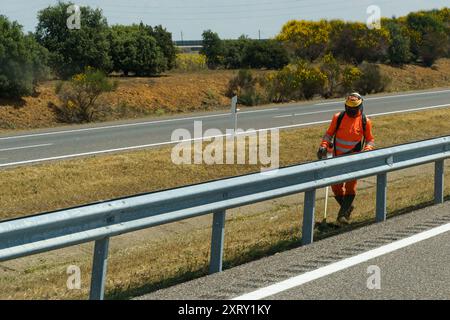 Astorga, Espagne - 5 juin 2023 : un travailleur en combinaison orange vif et casque coupe l'herbe le long d'une autoroute avec un outil. La route passe thro Banque D'Images