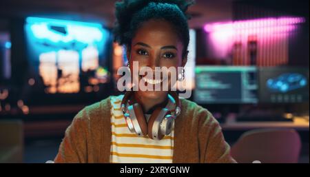 Portrait d'une jeune femme noire avec des écouteurs, regardant la caméra et souriant dans un bureau créatif. Belle femme diversifiée travaillant pour son agence de rêve, se sentant responsabilisée Banque D'Images
