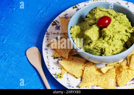 Guacamole dans un bol bleu avec une tomate cerise entourée de nachos qui sont sur une assiette décorée de fleurs sur un fond bleu. Banque D'Images