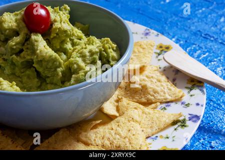 Guacamole dans un bol bleu avec une tomate cerise entourée de nachos qui sont sur une assiette décorée de fleurs avec une spatule en bois reposant dessus. Banque D'Images