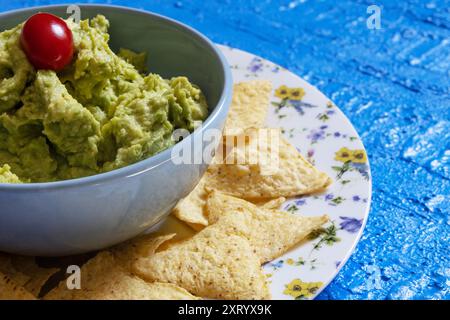 Guacamole dans un bol bleu avec une tomate cerise entourée de nachos sur une assiette décorée de fleurs. Banque D'Images