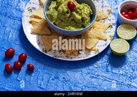 Belle table avec guacamole dans un bol entouré de nachos, tomates cerises et moitiés de citron vert. Banque D'Images