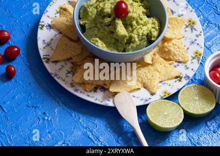 Belle table bleue avec guacamole dans un bol entourée de nachos, tomates cerises et moitiés de citron vert. Banque D'Images