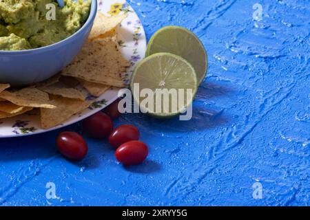 Partie d'un bol de guacamole entouré de nachos dans le coin supérieur gauche de l'image avec des tomates cerises et des moitiés de citron vert à côté. Banque D'Images