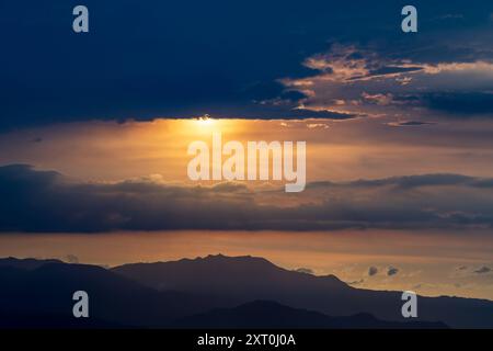 Un coucher de soleil doré partiellement obscurci par des nuages, avec le soleil apparaissant et disparaissant par intermittence, projetant des silhouettes des montagnes. Caoshan Ra Banque D'Images