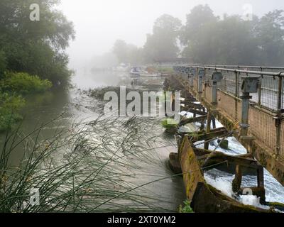 Abingdon-on-Thames prétend être la plus ancienne ville d'Angleterre. Et la Tamise traverse son cœur. Ici, nous avons son barrage, juste en amont de son pont médiéval, obstrué au point de rencontre de la rivière avec l'affluent Abbey Spring. Ce déversoir jouxte l'écluse d'Abingdon. Et c'est un matin d'été brumeux. Banque D'Images