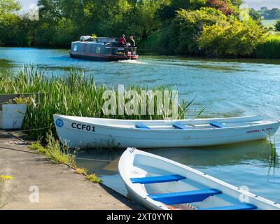 Fondé en 1921 et situé sur une belle partie de la Tamise dans l'Oxfordshire, Radley Boathouse sert Radley College et les amateurs d'aviron locaux depuis plus d'un siècle. Ici, nous voyons deux bateaux à rames amarrés par sa jetée, tôt par un beau matin d'été. Une péniche passe par là. Banque D'Images