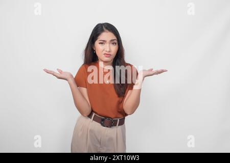 Belle jeune femme asiatique portant une chemise brune semble si confuse, isolée par un fond blanc. Banque D'Images