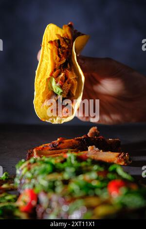 Une main de femme recadrée méconnaissable tenant un taco côtelé avec de la viande succulente et du basilic frais. Le taco est niché dans une tortilla de maïs jaune, près d'un g. Banque D'Images