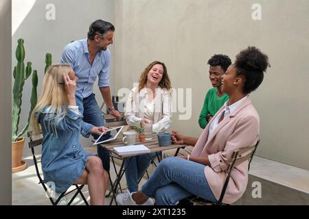 Des collègues multiraciaux positifs se rassemblent autour de la table sur la terrasse et rient tout en se refroidissant ensemble pendant le travail Banque D'Images
