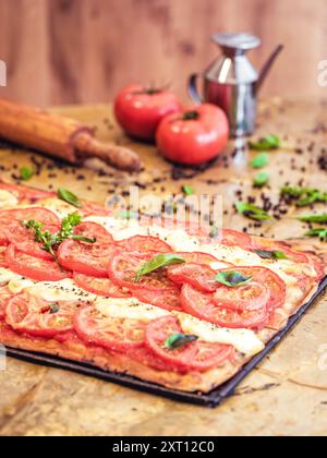 Tarte aux tomates fraîchement cuites et à la mozzarella garnie de basilic sur un comptoir de cuisine rustique, accompagnée de tomates, d'un rouleau à pâtisserie et d'une coloration Banque D'Images