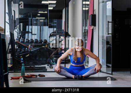 Une athlète en bleu effectue un étirement assis sur un tapis, se concentrant sur sa routine de flexibilité dans un cadre de gym Banque D'Images