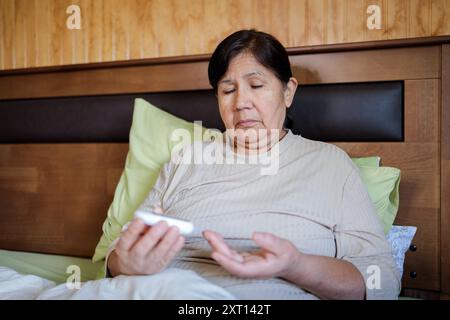 Une femme âgée est assise dans son lit en regardant préoccupée alors qu’elle vérifie son taux de sucre dans le sang avec un glucomètre, symbolisant la gestion de l’hypoglycémie Banque D'Images