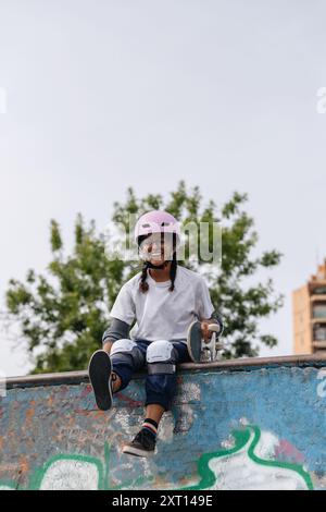 Corps complet de jeune afro-américaine gaie portant un t-shirt blanc avec casque et des coussins de sécurité avec planche à roulettes assis sur la rampe de skate dans le parc Banque D'Images