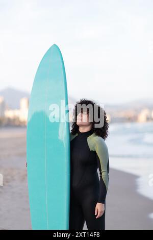 Jeune femme de surf attentionnée en combinaison avec planche de surf debout avec les yeux fermés sur le bord de mer lavé par la mer Banque D'Images