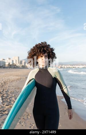 Jeune femme de surf attentionnés en combinaison avec planche de surf marchant en regardant loin sur le bord de mer lavé par la mer Banque D'Images