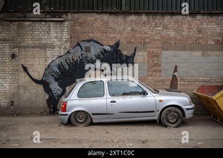 Londres, Royaume-Uni. 12 août 2024. Banksy Rhino art murale à Londres - la huitième œuvre d'art animalier en huit jours par le mystérieux artiste de rue. La dernière image montre l'animal montant une vieille Nissan Micra argentée - qui a un cône de circulation sur son capot, apparemment pour ressembler à un autre rhinocéros. La murale rhinocéros est située sur le côté d'un bâtiment de Westmoor Street à Charlton, au sud-est de Londres, près d'une cour utilisée par la société d'élimination des déchets RMS. Crédit : Guy Corbishley/Alamy Live News Banque D'Images