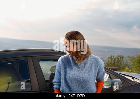 Une femme souriante dans un pull bleu se tient à côté d'une voiture avec une vue panoramique en arrière-plan. Bola02354 Copyright : xConnectxImagesx Banque D'Images