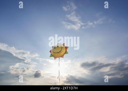 Un ballon souriant en forme de soleil flotte contre un ciel bleu vif avec des nuages moelleux dispersant la lumière du soleil. Bola02356 Copyright : xConnectxImagesx RECORD DA Banque D'Images