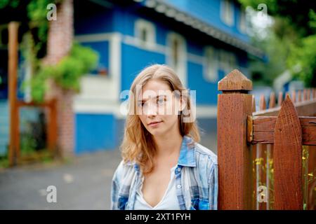 Une jeune femme avec une expression contemplative debout près d'une clôture en bois, avec une maison bleue en arrière-plan. Bola02501 Copyright : xConnectxImagesx Banque D'Images