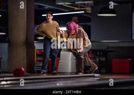 Groupe d'amis profitant d'un jeu amusant de bowling dans une piste de bowling moderne, en se concentrant sur la technique de l'individu que d'autres attendent et observent avec des sourires Banque D'Images