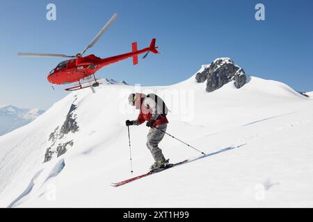 Skieur en rouge descendant une pente de montagne immaculée avec un hélicoptère rouge en vol en toile de fond. Phhi00037 Copyright : xConnectxImagesx Banque D'Images