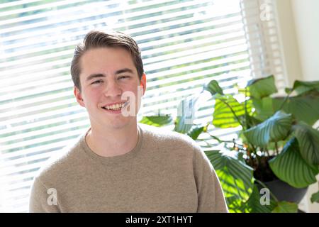 Portret van een jeune homme souriant millenial dans un pull décontracté debout à l'intérieur avec des stores de fenêtre et une plante verte en arrière-plan. Molenhoek Ned Banque D'Images